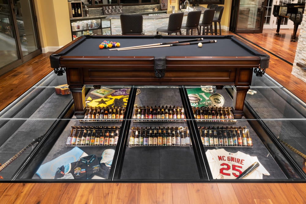 Pool table above glass floor panels showcasing neatly arranged cigar collections underneath.