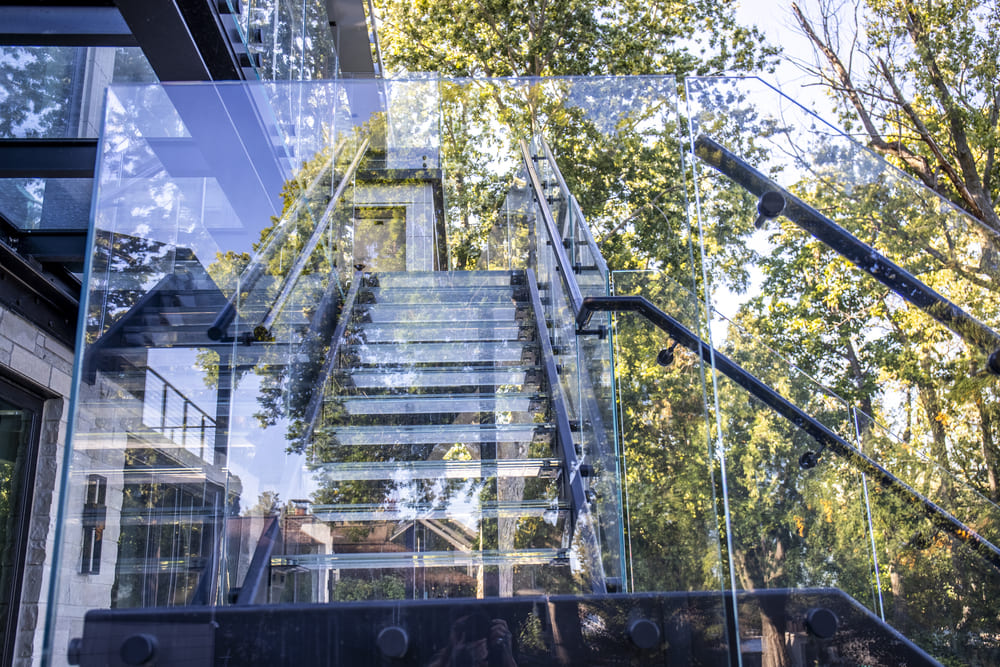 Outdoor glass staircase reflecting surrounding trees and modern architecture.