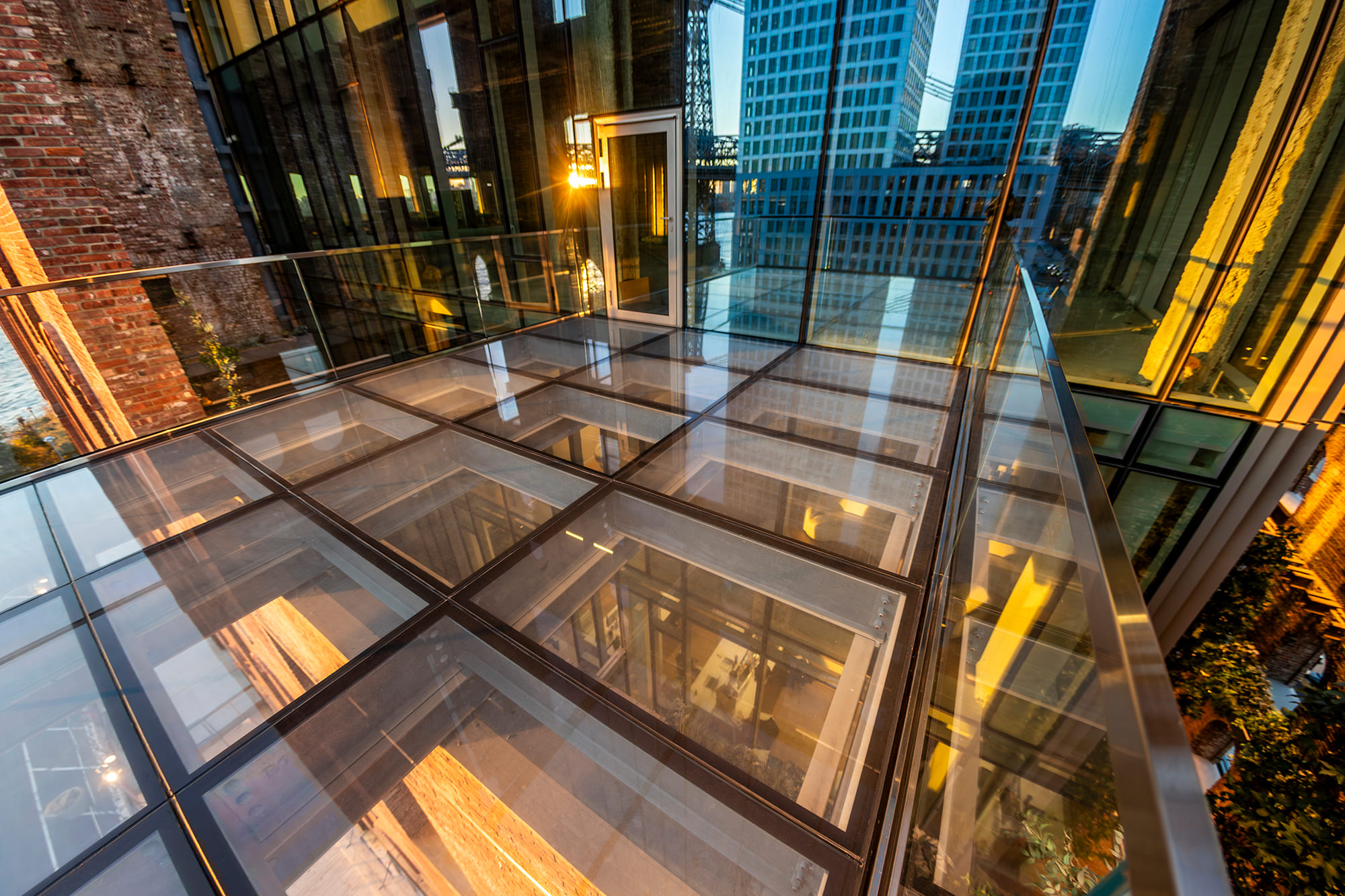 A glass floor walkway illuminated from below, surrounded by tall buildings and reflecting city lights.