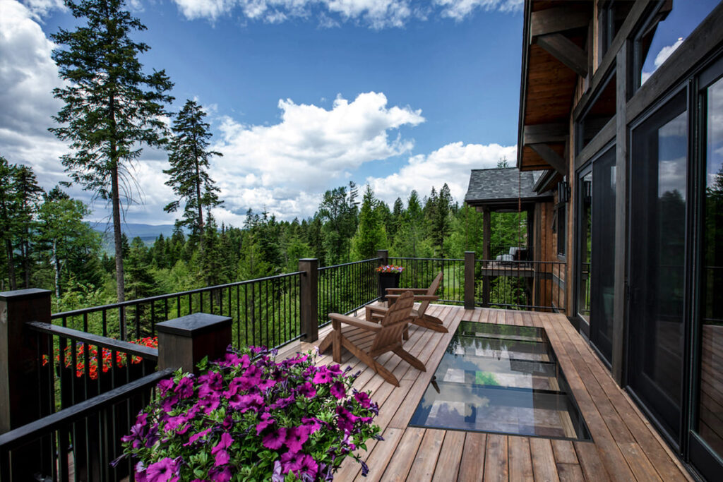 A wooden deck with a rectangular glass floor panel beside vibrant flowers, overlooking a lush forested landscape.