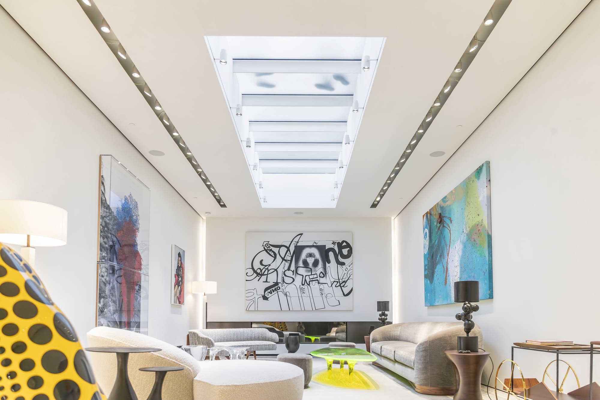 A contemporary living room with colorful artwork on the walls, white seating, decorative lamps, and a long skylight running down the center of the ceiling.