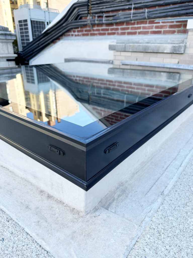 A close-up view of a modern flat glass skylight installed on a rooftop, featuring a black metal frame, reflective glazing, and surrounding concrete surface.