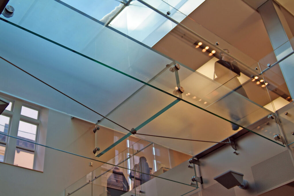 Interior view of a modern space with layered glass panels, metal fittings, and ceiling-mounted structural supports.