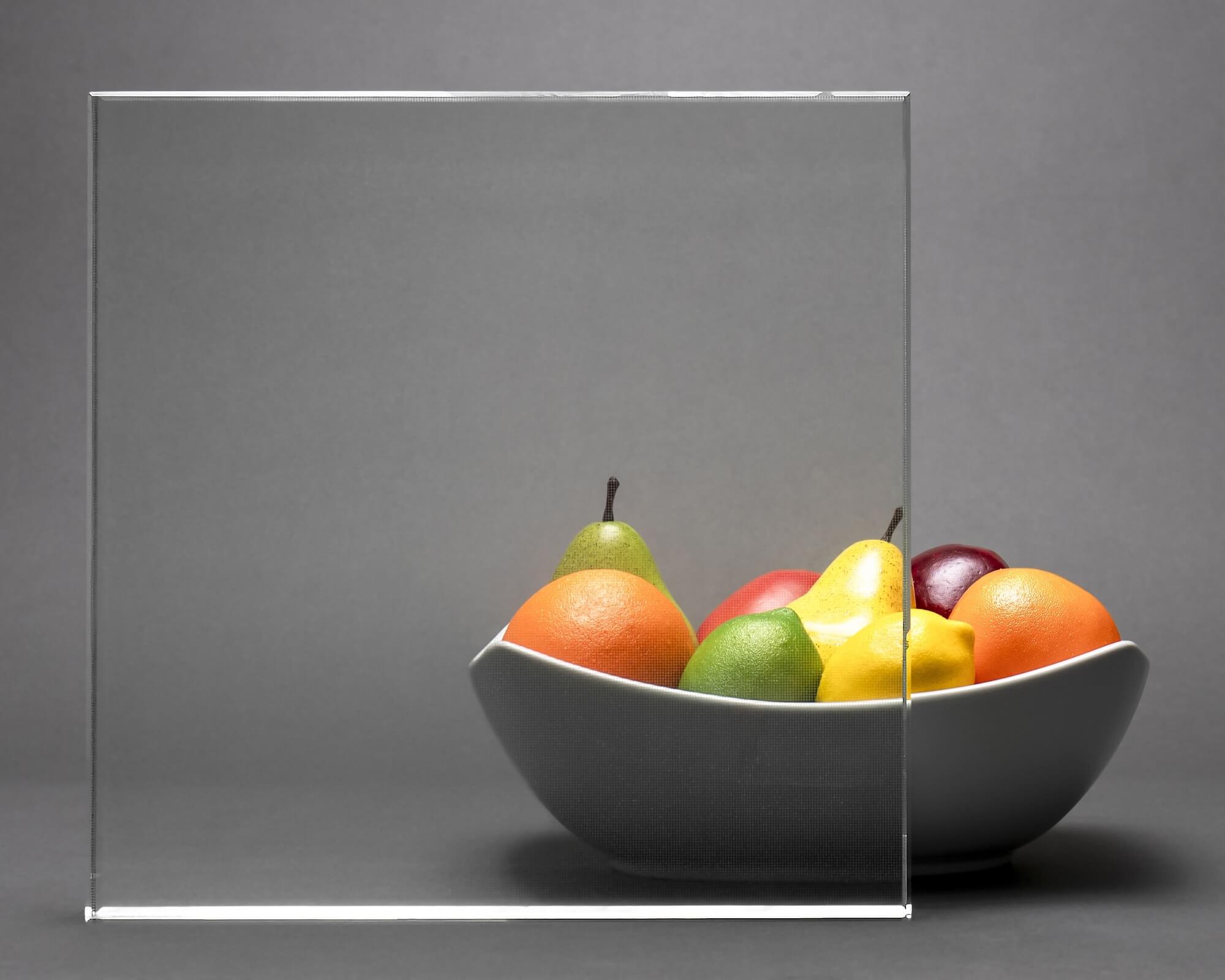 Minimalist still-life of a bowl of fruit displayed against a clear glass panel on a neutral background.