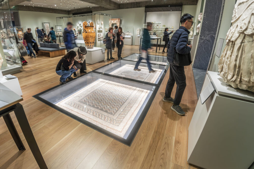 Visitors stand around a large, flat, illuminated display case set into a wooden museum floor, viewing a detailed artwork while others walk through the gallery.