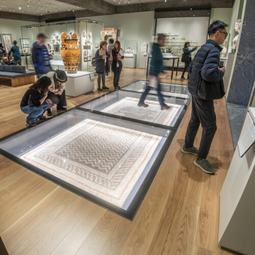 Visitors stand around a large, flat, illuminated display case set into a wooden museum floor, viewing a detailed artwork while others walk through the gallery.