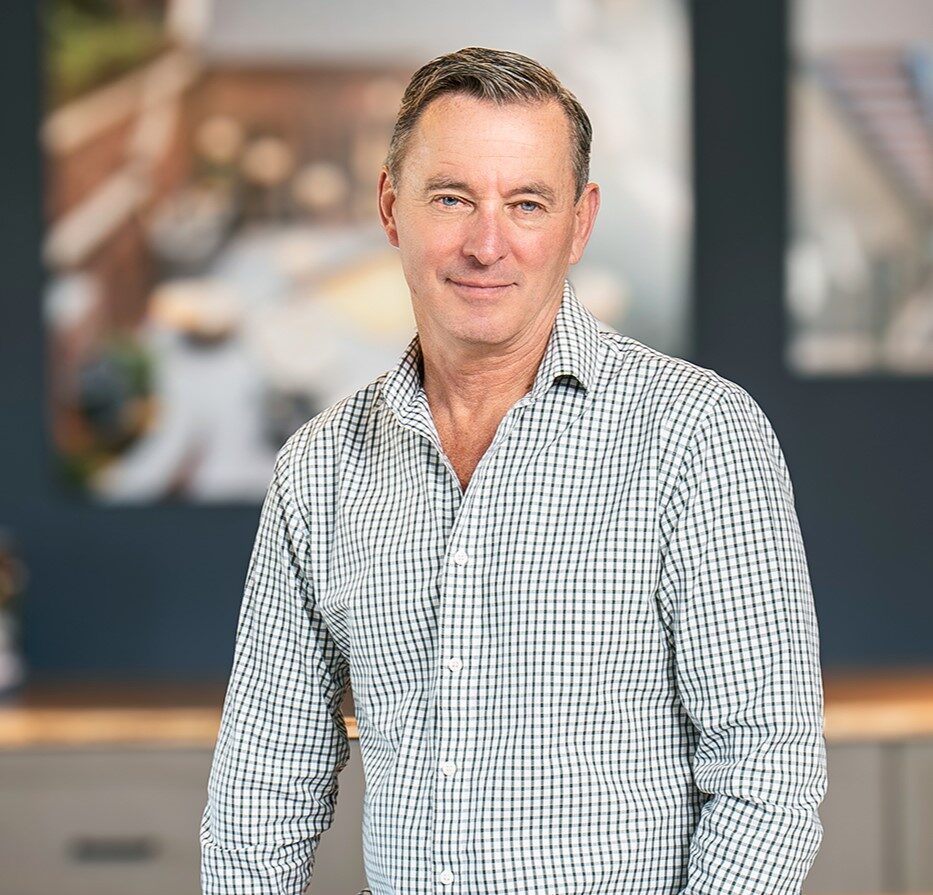 Man in a casual button-down shirt and jeans standing indoors and smiling, with a blurred office or workspace background.