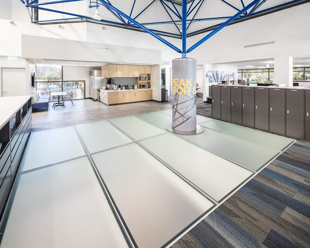 Modern interior space with large glass floor panels and exposed blue ceiling beams in a bright lobby or showroom.