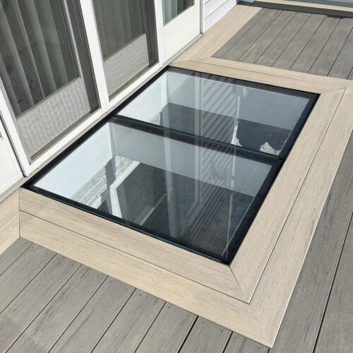 A two-panel walkable glass floor installed on an outdoor composite deck, located just outside a white sliding glass patio door.