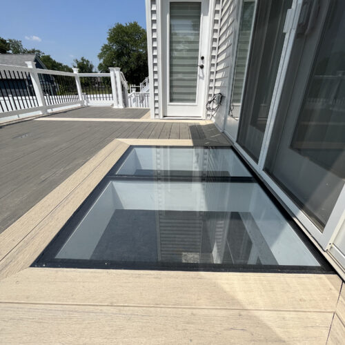 Two walkable glass deck panels set into composite decking beside patio doors, bringing natural light to the space below.