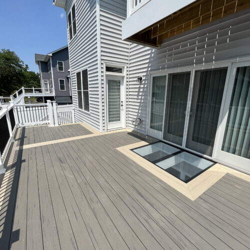 Exterior deck with two walkable glass floor panels framed by composite decking, positioned beside sliding patio doors to bring light below.