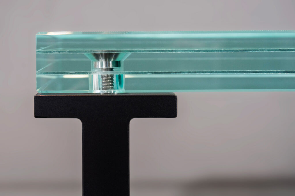 Close-up side view of a laminated glass stair tread with polished edges, a titanium embedded fastener, and a black aluminum support frame.
