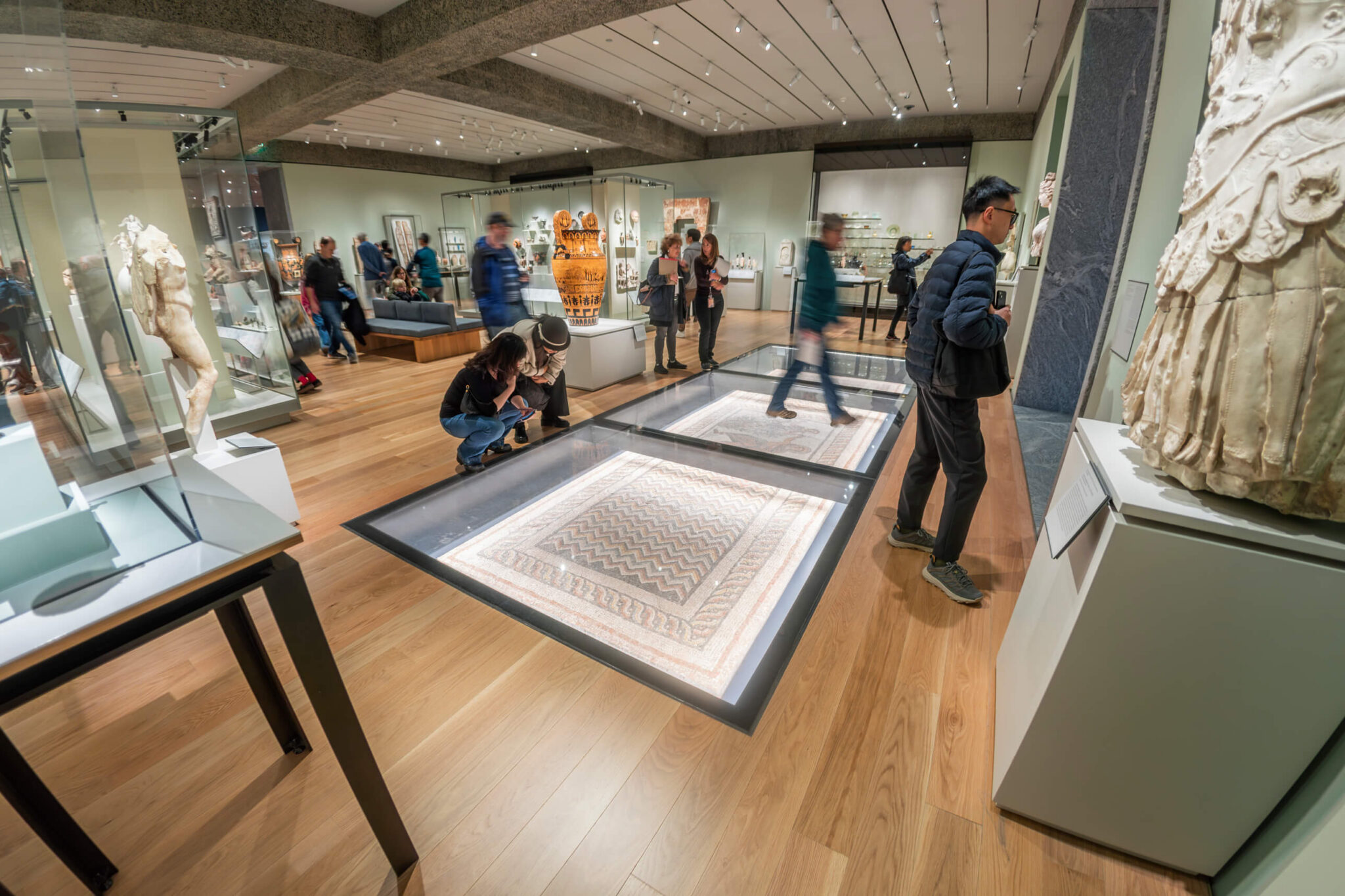 Museum visitors around a large illuminated glass floor display case set into a wooden gallery floor