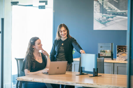 Two women in a conference room reviewing glass panel samples, with product displays and marketing visuals in the background.