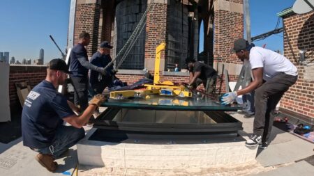 A group of workers installing a large glass skylight on a rooftop using lifting equipment, surrounded by brick structures and city buildings.