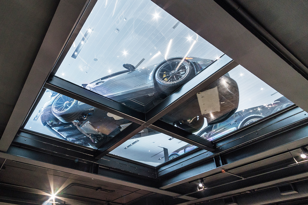 a shot of a silver supercar from the floor below, looking up through a drivable glass floor.