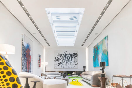A sleek modern living room with abstract artwork and neutral furniture is illuminated by a rectangular walkable glass skylight running the length of the ceiling.