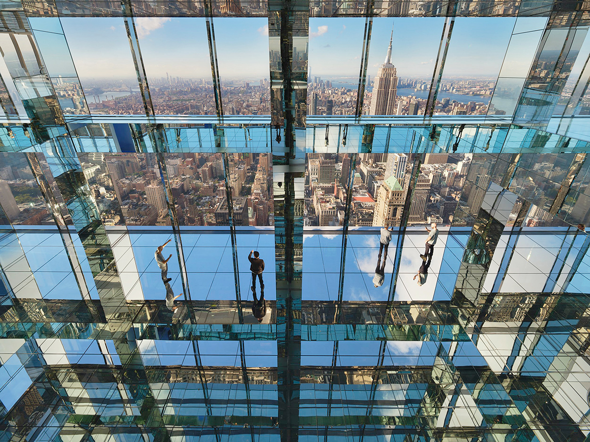 large glass windows at Summit One Vanderbilt in New York City, where we can see people walking inside as well as a reflection of NYC.