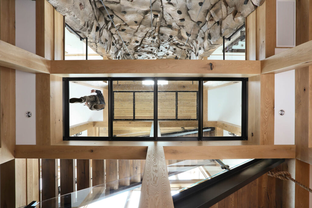 Looking up through a four-panel structural glass floor bridge to the climbing wall above, timber beams framing the recessed black steel frame
