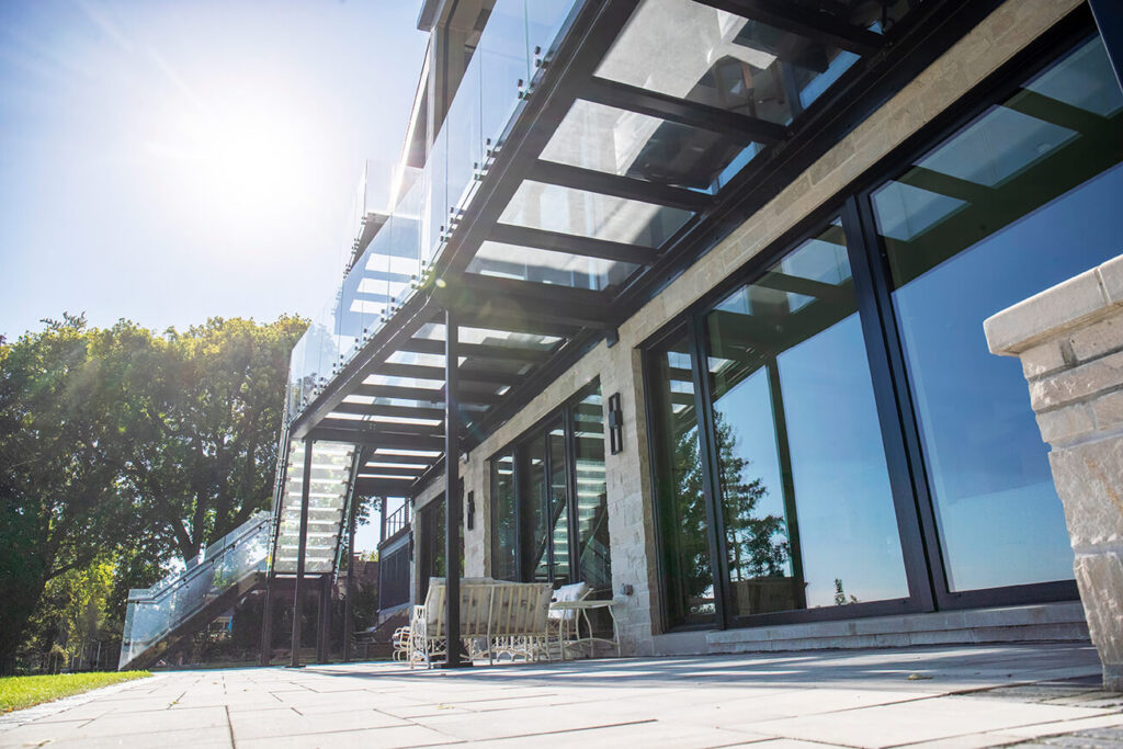 Looking up from the patio at the underside of a structural glass deck with black steel I-rafters and an exterior glass staircase