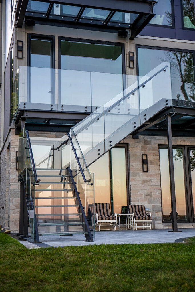 Exterior L-shaped glass staircase with laminated glass treads, glass railings and black steel stringers leading up to a structural glass deck