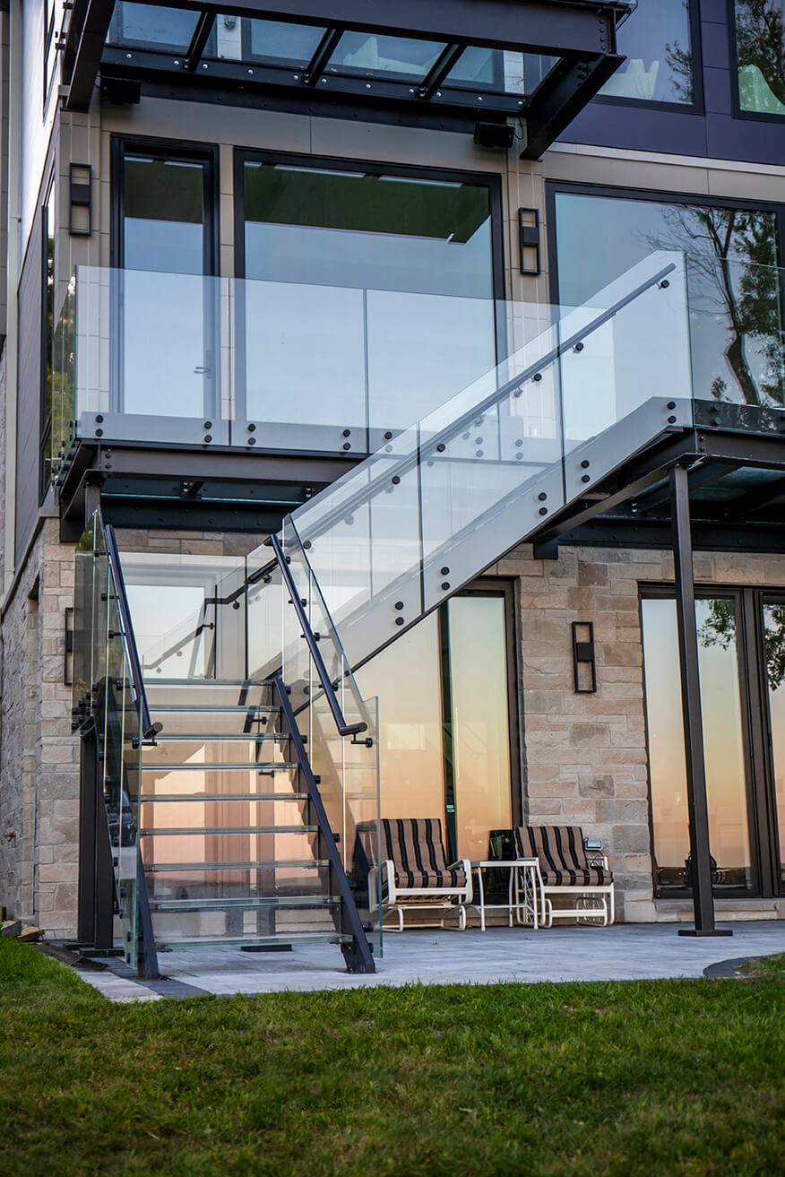 Exterior L-shaped glass staircase with laminated glass treads, glass railings and black steel stringers leading up to a structural glass deck