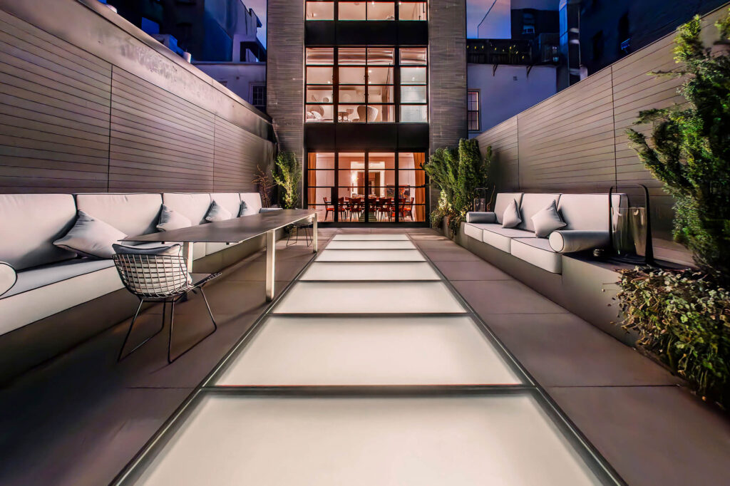 Rooftop terrace at night with a flush SkyFloor Series 2000 walkable skylight glowing between built-in seating, townhouse windows lit behind